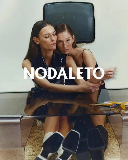 Two women in a close embrace seated beneath a glass table, with an old television set in the background, showcasing a modern fashion aesthetic.