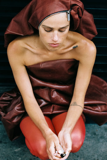 A young, light-skinned woman in a brown leather-like outfit kneels, her head bowed, hands together near her knees, against a dark background.