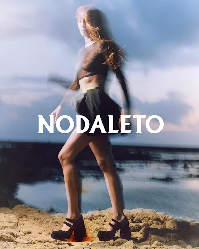 Fashion shot of a woman wearing a sheer top and black skirt with platform shoes, posing on a sandy beach with a cloudy sky backdrop.