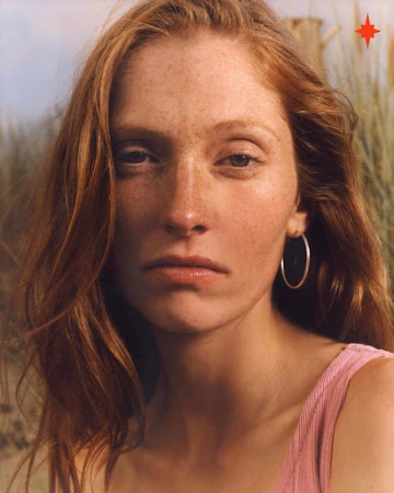 Close-up portrait of a woman with red hair, freckles, and silver hoop earrings against an outdoor grassy background.