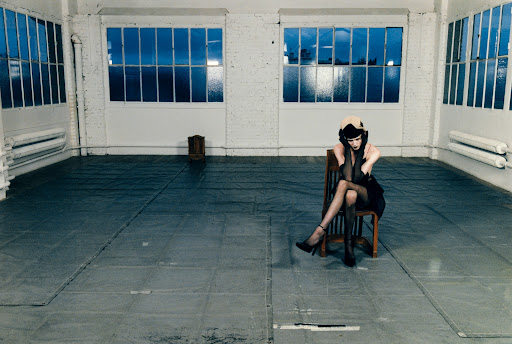 An adult female model in a dark dress sits in a chair in an empty room with large windows. The room has a gray floor.