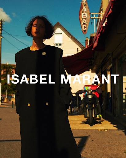 An adult female model stands on a city street wearing a black coat with visible text "ISABEL MARANT" in an outdoor fashion shot.