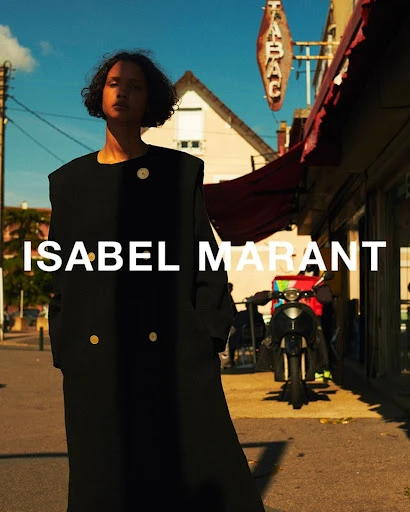 An adult female model stands on a city street wearing a black coat with visible text "ISABEL MARANT" in an outdoor fashion shot.