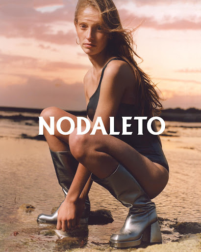 Fashion shot of a woman in a black swimsuit and silver boots squatting on wet sand in front of a pastel sunset sky by the ocean.
