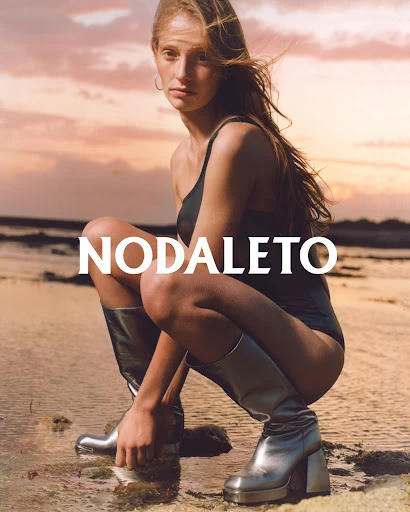 Fashion shot of a woman in a black swimsuit and silver boots squatting on wet sand in front of a pastel sunset sky by the ocean.
