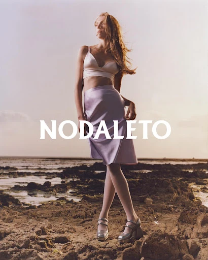 Fashion shot of a young woman in a top and skirt, posing on a rocky beach at sunset with designer logo text overlay, 'NODALETO'.