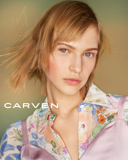 Close-up fashion portrait of a blonde adult female with fair skin, wearing a floral shirt and lavender top against a muted background.