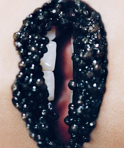 Close-up of a person's lips coated in black caviar-like beads with white teeth visible between the parted lips, beauty and art.