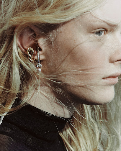 Close-up shot of a blonde woman with blue eyes wearing earrings; one is a silver hook with a pearl, the other is gold and twisted.