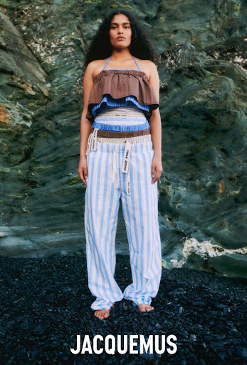 A woman posing in a halter top and striped pants stands on a rocky beach with a rocky cliff face in the background.