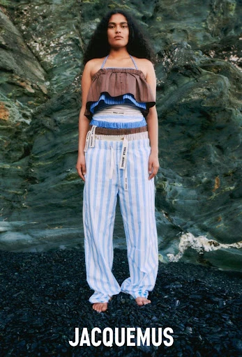 A woman posing in a halter top and striped pants stands on a rocky beach with a rocky cliff face in the background.