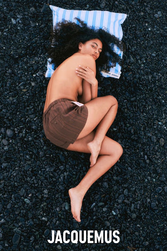 Top-down view of a woman with dark curly hair lying on a striped pillow on a black pebble beach wearing brown shorts.