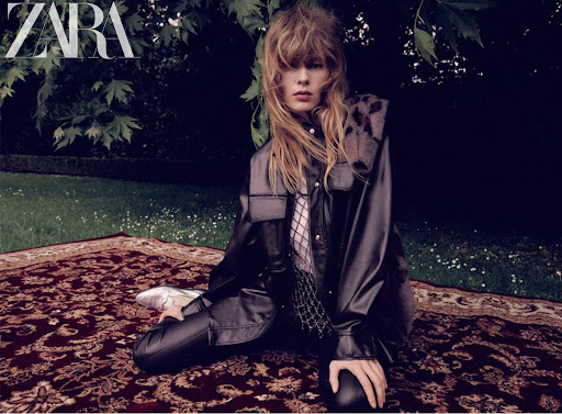 A blonde woman wearing a black leather jacket and pants sits on a patterned red rug in a dark outdoor setting with green foliage.