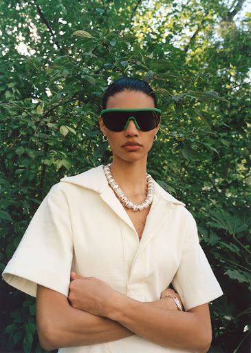 A woman poses outdoors wearing sunglasses, a light beige jumpsuit, and a shell necklace against a blurred green leafy background.