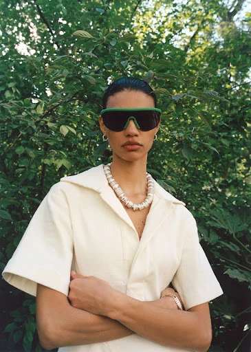 A woman poses outdoors wearing sunglasses, a light beige jumpsuit, and a shell necklace against a blurred green leafy background.