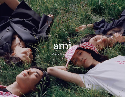 Group of four young adults laying in green grass, viewed from above, promoting the clothing brand Ami Alexandre Mattiussi.
