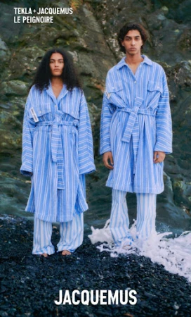Two models wearing blue and white striped robes and pants standing on a rocky shore with sea foam.