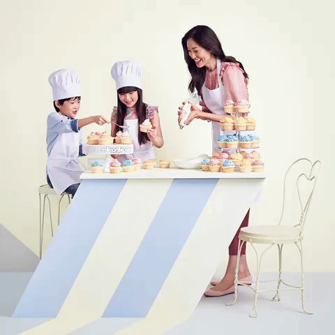 A happy family of three, including a woman and two children, baking cupcakes together in a light-filled indoor setting.