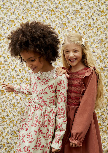 Two cute girls with an African and a Caucasian girl pose side by side in their fashionable dresses against a floral backdrop.