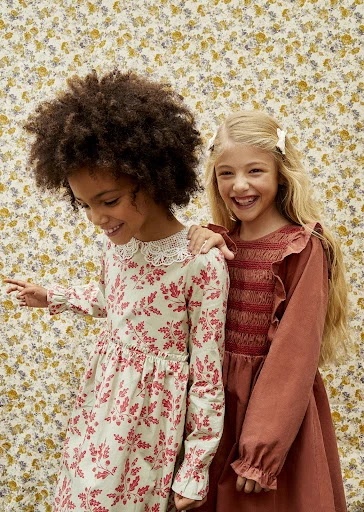 Two cute girls with an African and a Caucasian girl pose side by side in their fashionable dresses against a floral backdrop.