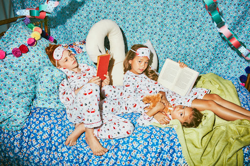 Three young girls wearing matching pajamas and sleep masks are lying on a bed with a blue floral pattern reading books.