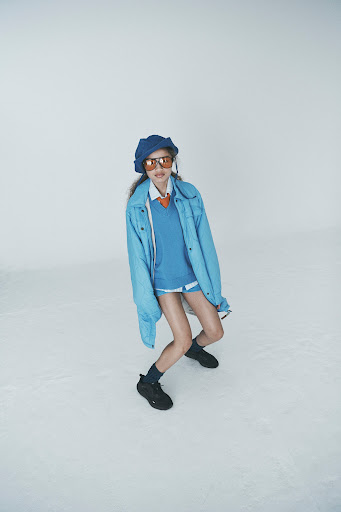 Stylish young woman with sunglasses, blue beret, denim jacket, and black sneakers posing in a light, airy studio setting.