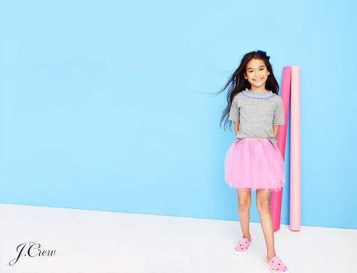 A young girl in a grey t-shirt and pink tutu stands against a light blue backdrop in a studio shot. She is smiling.
