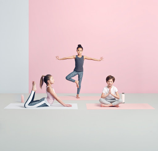 Three children practice yoga poses on mats in a studio setting with a pink and white wall background, promoting fitness and flexibility.