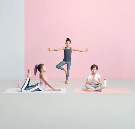 Three children practice yoga poses on mats in a studio setting with a pink and white wall background, promoting fitness and flexibility.