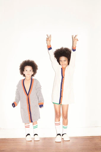 Two children with afro hairstyles wearing matching vintage-style cardigan sweaters, knee-high socks, and white shoes in a white studio.