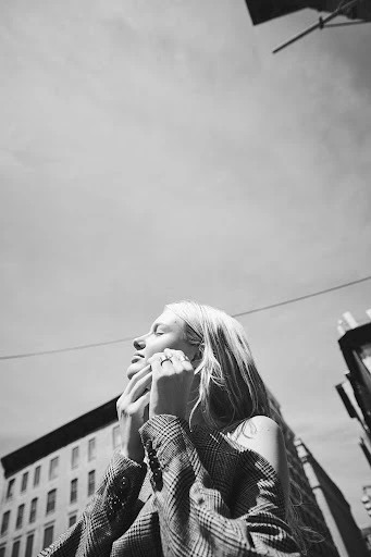 Low angle black and white portrait of a blonde woman in a blazer, looking up with closed eyes, in an urban setting with buildings.