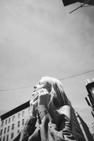 Low angle black and white portrait of a blonde woman in a blazer, looking up with closed eyes, in an urban setting with buildings.
