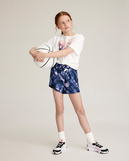 Red-haired tween girl in tie-dye shorts holds a basketball in a studio setting, wearing a white t-shirt and athletic shoes.