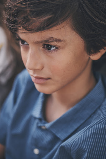 Portrait of a young boy with dark hair wearing a blue collared shirt, looking to the side in a close-up shot, captured in natural light.