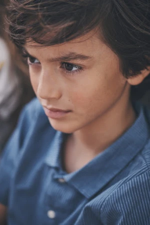 Portrait of a young boy with dark hair wearing a blue collared shirt, looking to the side in a close-up shot, captured in natural light.