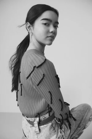 Portrait of a young woman with long dark hair in a ponytail, wearing a sweater and jeans, gazing over her shoulder in a photo studio.