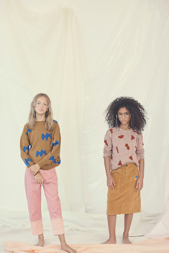Two young girls model patterned knit sweaters and casual bottoms in a brightly lit, neutral studio environment, showcasing youth fashion.