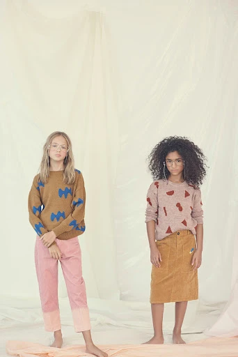 Two young girls model patterned knit sweaters and casual bottoms in a brightly lit, neutral studio environment, showcasing youth fashion.