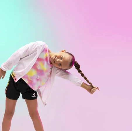A young girl in athleisure wear poses against a pink and turquoise gradient backdrop, smiling with her hair in a long braid.