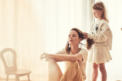 Woman seated on the floor with a young child styling her hair near a bright window with a white chair in the background