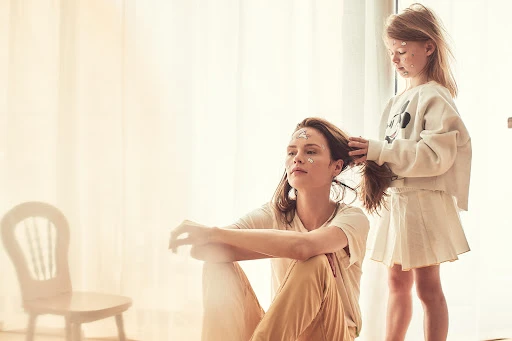 Woman seated on the floor with a young child styling her hair near a bright window with a white chair in the background