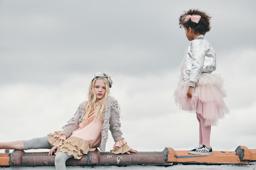 Two young girls in fashionable dresses pose on a rooftop with a cloudy sky in the background, showcasing children's fashion and outdoor setting.