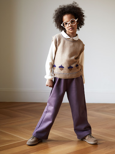 Child standing indoors wearing an acorn-themed sweater vest, long-sleeve shirt, wide-legged purple pants, glasses, and tan shoes on a wood floor.