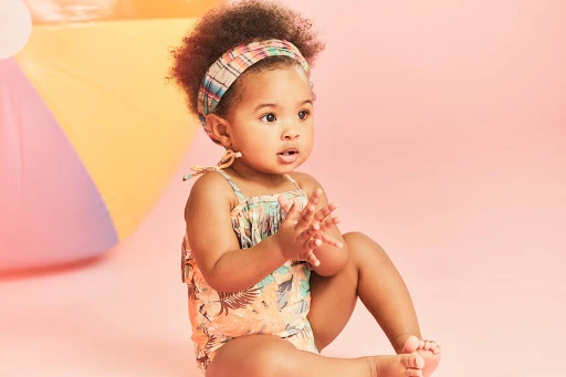 A young child with a patterned headband and floral swimsuit sits in front of a pink background with an out-of-focus beach ball.