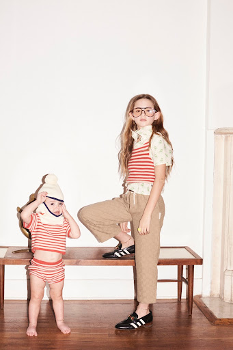 Two children pose indoors against a white wall. A younger child in striped shirt and diaper stands next to an older child.