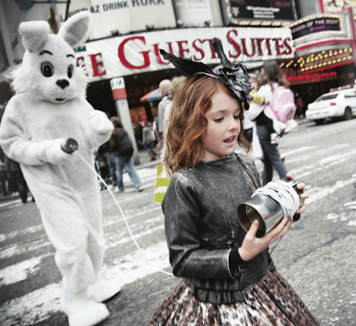 Young girl with a bunny mascot using tin can phones outdoors in a city. Building signage is visible in the background.