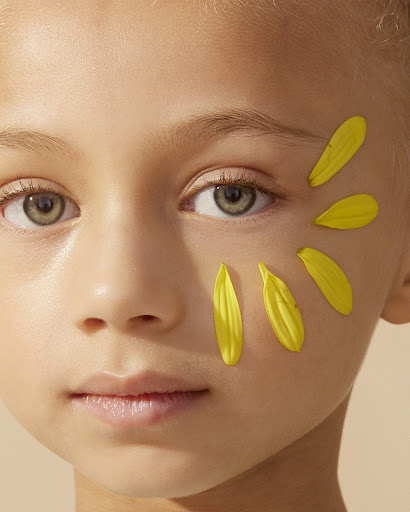 Close-up portrait of a fair-skinned child with yellow flower petals near their eye, set against a soft beige background in eye-level shot.