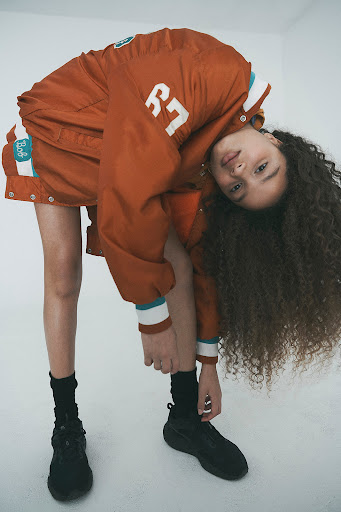 Model with long curly hair wearing an orange varsity jacket bending upside down against a white background