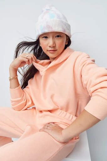 Child model poses indoors wearing a pastel-colored beanie and peach sweatsuit against a white background in a medium shot.