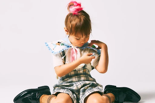 A young child with pink-tipped hair sits on a white surface wearing a plaid romper and a patterned collar, looking down at an object.